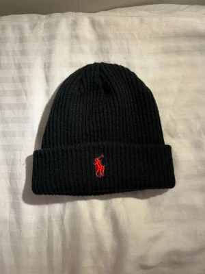 Svart stickad mössa från Polo Ralph Lauren - Säljer en svart ribbstickad mössa från Polo Ralph Lauren med röd broderad logga framtill. Mössan är klassisk med uppvikt kant och är gjord i mjuk ull som håller dig varm under kyliga dagar. Perfekt accessoar för att lyfta din vinterstil. Nypris 1100kr.