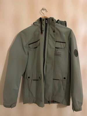 Grön vindjacka med rutig huva - Snygg grön vindjacka från Mountain Top med svart dragkedja och coola fickor framtill. Jackan har huva med rutigt foder och svarta detaljer. Perfekt för dig som gillar streetstyle och vill ha något praktiskt med lite edge.