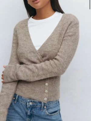 Beige omlottkofta från Gina  - Säljer en beige omlottkofta från & Other Stories med v-ringning och långa ärmar. Koftan har en croppad passform och stängs med fyra knappar framtill. Materialet är mjukt och stickat, perfekt för lager på lager under höst och vinter.