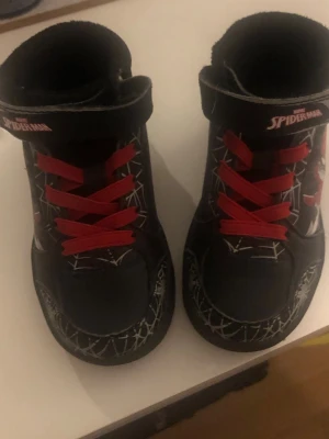 Svarta Spiderman sneakers med röd snörning - Säljer ett par coola svarta sneakers med Spiderman-tryck och röda snören. Skorna har vita spindelnätsmönster och kardborreband upptill. Rund tå och platt sula, tillverkade i syntetmaterial. Perfekta för dig som gillar superhjältar!