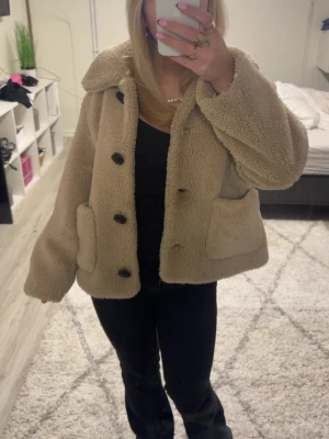 Beige teddyjacka från Zara, S - Supermysig teddyjacka från Zara i beige färg med stora svarta knappar och två rejäla fickor framtill. Jackan har en loose passform och är tillverkad i fluffigt syntetmaterial som håller dig varm. Perfekt för dig som gillar chill och comfy vibes.