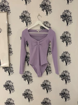 Ljuslila ribbad body med rosett - Supersnygg ljuslila body från H&M i ribbad bomullsmix. Modellen har lång ärm, söt rosettdetalj framtill och en djup, fyrkantig ringning. Perfekt att styla med jeans eller kjol för en trendig look.