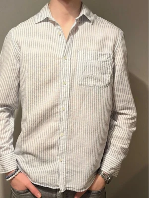 Vit och blå randig skjorta från Zara - Säljer en fräsch vit skjorta med tunna blå ränder från Zara. Skjortan har klassisk krage, lång ärm och en bröstficka. Tillverkad i lätt och luftig bomull, perfekt för dig som gillar stilrena och enkla plagg. Knappar framtill och relaxed passform.