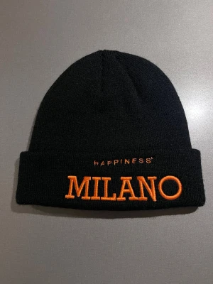 Svart mössa från Happiness Milano - Svart mössa från Happiness med brodyr i orange framtill och en unik orange detalj på Duomo på baksidan. Tillverkad i Italien och köpt i Happiness butiken i Milano. Skön och varm. Passar M/L. Mycket bra skick, som ny! Säljs på flera sidor, först till kvarn. 