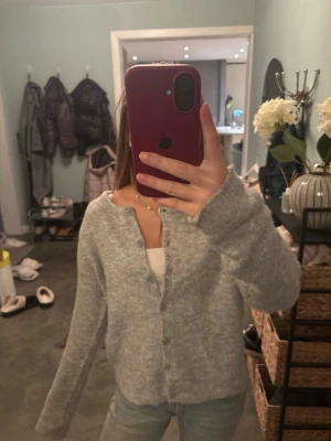 Grå stickad kofta med knappar - Mysig grå stickad kofta med rund hals och knappar framtill. Koftan har långärmade ärmar och en loose passform som gör den perfekt att slänga på över en t-shirt eller linne. Enkel och stilren design som passar till jeans eller kjol. Köpt utomlands