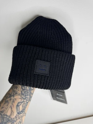 Svart stickad mössa Acne Studios - Säljer en svart, ribbstickad mössa från Acne Studios med den ikoniska ansiktsloggan framtill. Mössan är tjock och har bred uppvikt kant, perfekt för kalla dagar. Materialet är mjukt och värmande ull som känns lyxigt och trendigt.
