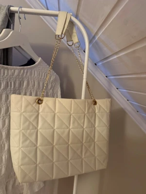Vit axelväska  - En stilren axelväska i vit färg med guldfärgade kedjehandtag. Väskan har ett rutmönster. Perfekt för dig som gillar en clean och chic look med lite lyxig känsla.