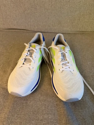 Adidas Supernova vita löparskor - Fräscha Adidas Supernova löparskor i vitt med neon-gula och blå detaljer. Skorna har ventilerande mesh-överdel, klassisk snörning och platt sula med Dreamstrike-dämpning. Perfekta för träning och löpning, snygg design med ikoniska ränder på sidan.