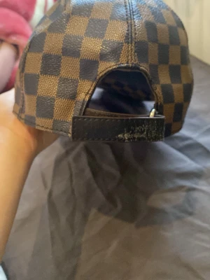 Rutig Louis Vuitton keps i skinn - Säljer en snygg keps från Louis Vuitton med klassiskt rutigt mönster i brunt och svart. Kepsen är tillverkad i skinn och har justerbar rem baktill. Märket syns både på insidan och på skärmen. Perfekt för dig som gillar exklusiva accessoarer.