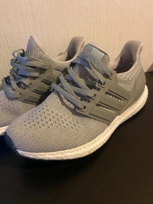 Adidas Ultraboost grå  - Säljer ett par Adidas Ultraboost sneakers i ljusgrå färg med stickad ovandel och vita Boost-sulor. Skorna har snörning och klassiska tre ränder på sidan i plast. Perfekta för dig som gillar stilrena och bekväma sneakers med modern design.