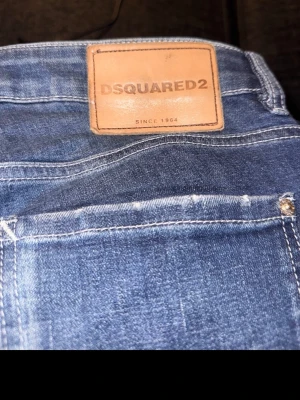 Blå jeans från Dsquared2 - Säljer ett par blå jeans från Dsquared2 med klassisk femficksdesign och snygga slitningar framtill. Jeansen har en normal passform och är tillverkade i jeansmaterial med tydliga kontrastsömmar och läderpatch med logga bak i midjan. 