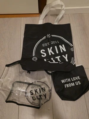 Skin City shoppingväska och necessär - Säljer ett set med Skin City-väskor: en svart shoppingväska i syntet med vita handtag och tryck, en transparent necessär med svart kant och tryck, samt en mindre svart väska med vit text. Perfekt för att organisera dina grejer och ha med på språng. Den genomskinliga väskan har svarta missfärgningar från att ha legat på den svarta påsen.