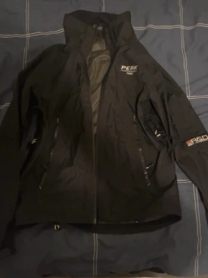 Svart vindjacka Peak Performance S - Svart vindjacka från Peak Performance med GORE-TEX-material och coola R&D-detaljer på ärmen. Jackan har dragkedja framtill, hög krage och flera praktiska fickor. Perfekt för dig som gillar stil och funktion när du är ute.