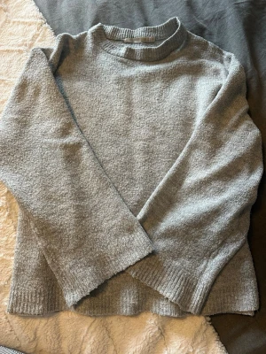 Grå stickad tröja från 157 - Mysig grå stickad tröja från 157 med ribbad krage, ärmslut och nederkant. Tröjan har en enkel, klassisk design med lång ärm och lite högre hals. Perfekt för lager-på-lager under kyliga dagar.