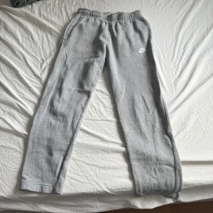 Grå mjukisbyxor från Nike - Grå mjukisbyxor från Nike med elastisk midja och sidofickor. Byxorna har en klassisk rak passform och är tillverkade i mjukt bomullsmaterial. Nike-logga i vitt på ena benet ger en sportig vibe. Perfekta för chill eller träning.