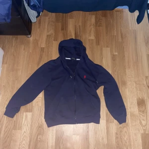 Marinblå hoodie från Polo Ralph Lauren - Passar XS/S Marinblå hoodie från Polo Ralph Lauren med dragkedja, huva och snörning. Klassisk röd logga broderad på bröstet och känguruficka framtill. Tillverkad i mjuk bomull, perfekt för chill dagar eller när du vill ha en clean streetstyle.