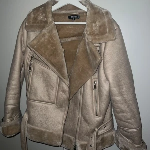 Beige pilotjacka från Missguided - Snygg beige pilotjacka från Missguided med mjukt foder, bred krage och detaljer i fuskpäls. Jackan har dragkedjor, bälte i midjan och uppvikta ärmslut. Jackan är väldigt varm