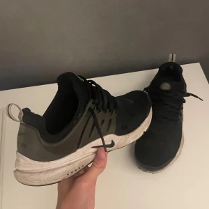 Nike Presto svarta sneakers - Svarta Nike Presto sneakers med vita sulor och grå detaljer. Skorna har snörning, rund tå och är tillverkade i syntetmaterial och textil. Perfekta för dig som gillar en sportig och clean stil. Swoosh-logga på sidan och AIR-detalj bak.