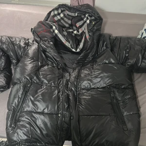 Svart pufferjacka från Burberry - Säljer en svart pufferjacka från Burberry med glansig finish, huva och klassiskt rutigt foder. Jackan har dragkedja framtill, stora fickor med dragkedja och en patch med Burberry London England på bröstet. Perfekt för kalla dagar och riktigt snygg streetstil.