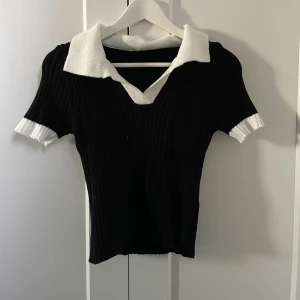 Svart ribbad topp med vit krage - Snygg svart ribbad topp med vit krage och vita detaljer vid ärmsluten. Toppen har v-ringning och korta ärmar.
