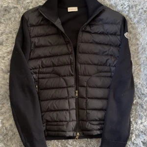 Svart (2022) cardigan jacka från Moncler M - Svart cardigan jacka från Moncler med quiltad, dunfylld front och stickade ärmar. Jackan har dragkedja framtill, Moncler-logga på ärmen och en clean, sportig look. Perfekt för dig som gillar stilren design och premiumkänsla.