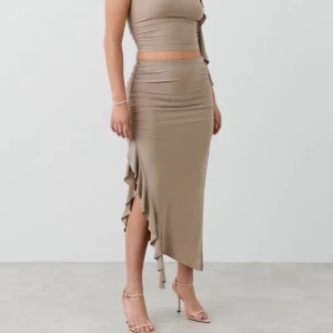 Beige långkjol med volang och slits - Snygg beige långkjol med tight passform och hög midja. Kjolen har en cool slits på sidan med volangdetalj som ger extra edge. Materialet är stretchigt och följsamt, perfekt för dig som gillar en stilren men ändå trendig look. Stolekn på den är ett M men passar även nån som har storlek S 