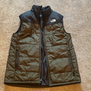 Svart dunväst från The North Face - Snygg svart dunväst från The North Face med hög krage och två dragkedjefickor framtill. Västen har quiltad design och logga på bröstet. Perfekt för lager-på-lager och håller dig varm under kyliga dagar.