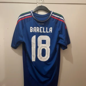 Italien Barella #18 Adidas matchtröja - Italiens landslagströja från Adidas med Barella och nummer 18 på ryggen. Klassisk blå färg med vita detaljer, trikolorfärgade ränder på axlarna och broderat landslagsmärke. Tillverkad i lätt och ventilerande AEROREADY-material, perfekt för fotboll eller supporterliv. Storlek L men har personligen M och den passar lika fint. 