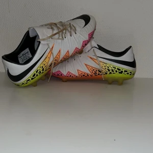 Nike Hypervenom fotbollsskor - Säljer ett par Nike Hypervenom fotbollsskor i vitt med svarta detaljer och coola grafiska mönster i neonrosa, orange och gul. Skorna har snörning och platt sula med dobbar för konstgräs. Perfekta för dig som vill sticka ut på planen! Storlek 44, passar 43/44 Skick 8/10, knappt använda.