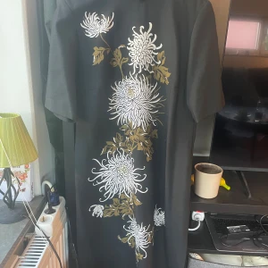 Svart långklänning med blommor - Svart långklänning från Treidun Design med stora vita och olivgröna blommor tryckta längs hela framsidan. Klänningen har korta ärmar och rak passform, perfekt för dig som vill ha något unikt och stilrent. Materialet känns mjukt och svalt.