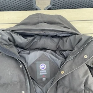 Svart dunjacka från Canada Goose - Säljer en svart dunjacka från Canada Goose med huva och klassisk logga på ärmen. Jackan är tjockt vadderad, har dragkedja och knappar framtill samt flera fickor. Perfekt för kalla vinterdagar och riktigt snygg streetstil.