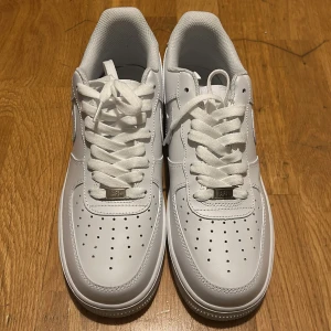 Nike Air Force 1 helvita sneakers - Klassiska Nike Air Force 1 sneakers i helvitt skinn med vit sula och snörning. Modellen har en låg profil, rund tå och perforerade detaljer på tån för extra ventilation. Perfekt för dig som vill ha en clean och tidlös look.