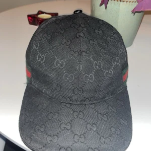 Svart Gucci keps med GG-mönster - Svart keps från Gucci med klassiskt GG-monogram över hela ytan. Kepsen har gröna och röda ränder på sidan, typiska för märket. Tillverkad i bomull och polyester, med justerbar rem baktill för perfekt passform. Snygg och stilren accessoar för dig som gillar lyxiga detaljer. 