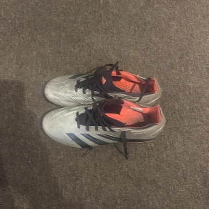 Adidas Predator silver fotbollsskor - Säljer ett par Adidas Predator fotbollsskor i silver med svarta detaljer och svarta snören. Skorna har orange insida och orange detaljer på sulan. Perfekta för dig som vill sticka ut på planen med snygg design och bra grepp.