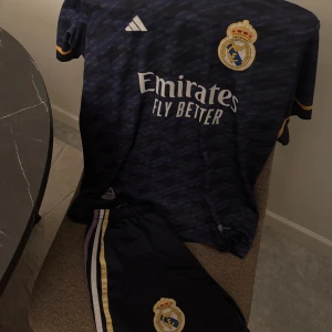 Real Madrid blå matchtröja Adidas - Säljer en officiell Real Madrid fotbollströja med shorts från Adidas i mörkblått med subtilt mönster. Tröjan har korta ärmar, gulddetaljer vid ärmslut och klubbens emblem på bröstet. Materialet är lätt och andas, perfekt för träning eller match. 5 bellingham på baksidan
