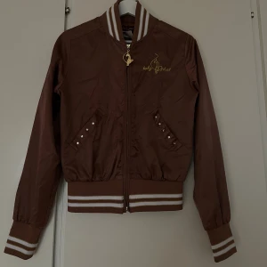 Brun bomberjacka från Baby Phat - Snygg brun bomberjacka från y2k-märket Baby Phat med guldbroderad kattlogga fram och bak. Jackan har ribbade muddar med vita och guldiga ränder, glittriga stenar vid fickorna och dragkedja med guldfärgad logga. Tunn jacka - mer som en college jacka eller kofta! 