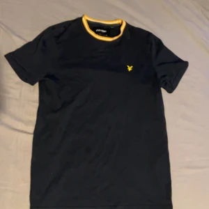 Svart t-shirt från Lyle & Scott - Svart t-shirt från Lyle & Scott med gul logga på bröstet och snygg ribbad krage med vita och gula ränder. Slim fit-modell i mjuk bomull, perfekt för en clean och stilren look.