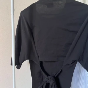 Svart topp med knyt från Nelly - Svart topp från Nelly med trekvartsärm och rund halsringning. Toppen har en snygg knyt-detalj i ryggen som ger en cool twist. Tillverkad i mjuk bomull, perfekt för dig som gillar stilrena och enkla plagg med en extra detalj.