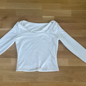 Vit långärmad topp med rundad urringning - En stilren vit långärmad topp med rundad, lite djupare urringning. Toppens passform är figurnära och den är tillverkad i ett mjukt, stretchigt material som känns skönt mot huden. Perfekt att matcha med jeans eller kjol för en clean look.