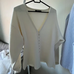 Vit kofta med v-ringning från Gina Tricot - Säljer en stilren vit kofta från Gina Tricot med djup v-ringning och knappar framtill. Koftan har lång ärm och är tillverkad i ett mjukt material som känns skönt mot huden. Perfekt att slänga på sig när det är lite kyligare ute.