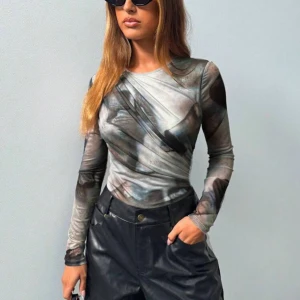 Grå mönstrad långärmad bodysuit - Säljer en riktigt snygg långärmad bodysuit med abstrakt mönster i olika grå och mörka toner. Bodyn har rund hals och coola draperade detaljer framtill som ger en unik look. Materialet är stretchigt och känns skönt mot huden. Perfekt att styla med jeans eller skinnbyxor.