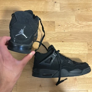 Svarta Nike Air Jordan 4 sneakers - Säljer ett par helsvarta Nike Air Jordan 4 sneakers med klassisk siluett och Jumpman-logga i silver på häl, plös och sula. Skorna har snörning, meshpaneler och detaljer i syntet och mocka för en riktigt clean look. Perfekta för dig som gillar streetwear och vill sticka ut.