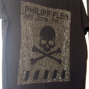  Philipp Plein t-shirt  - Svart t-shirt från Philipp Plein med glittrande detaljer framtill och stor dödskalle- och korsben-tryck. Klassisk passform, korta ärmar och rund hals. Märkeslogga i nacken och exklusiv känsla. Perfekt för dig som gillar edgy och lyxig streetstyle.