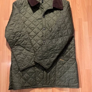 Olivgrön quiltad jacka från Barbour - Snygg olivgrön quiltad jacka från Barbour med brunt manchestertyg på kragen. Jackan har tryckknappar framtill, stora fickor och klassisk Barbour-logga broderad på ena fickan. Perfekt för höst och vår, riktigt stilren och tidlös design.