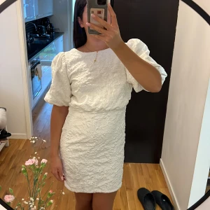 Vit kort klänning från Bubbleroom - Superfin vit kort klänning från Bubbleroom med puffärmar, strukturmönster och öppen rygg med stor rosett. Klänningen har en figurnära passform och är tillverkad i ett glansigt material som känns lyxigt. Perfekt för dig som vill sticka ut! Endast använd på student dagen.🩷