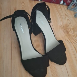 Svarta klackskor med öppen tå - Stilrena svarta klackskor från NLY Shoes med bred rem över tårna och justerbart ankelband. Skorna har öppen tå och en stadig blockklack, perfekta för att lyfta din outfit. Materialet är mockaliknande och ger en lyxig känsla. Priset jag köpte dem för står under. 