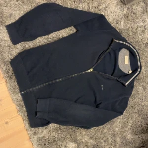 Gant kofta - Säljer en mörkblå kofta från Gant med hel dragkedja och diskret logga på bröstet. Tröjan har ribbade muddar, ståkrage med vit rand och är tillverkad i bomull. Ny pris 1700