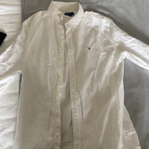 Vit skjorta från Gant, 170 cm - Klassisk vit skjorta från Gant med broderad logga på bröstet. Skjortan har button-down krage, långa ärmar och knäppning framtill. Tillverkad i bomull för en skön och stilren känsla. Perfekt för dig som gillar en clean och tidlös look. Storlek: 170cm, skick: 8/10