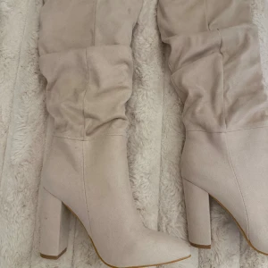 Beige höga stövlar med klack - Säljer ett par snygga höga stövlar i beige mocka-look med spetsig tå och rejäl blockklack. Stövlarna har en slouchy passform och är perfekta för att lyfta vilken outfit som helst. Supersnygga till både jeans och kjol!
