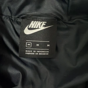 Svart pufferjacka från Nike med huva - Svart pufferjacka från Nike med vit logga på bröstet, justerbar huva och dragkedja framtill. Jackan har quiltad design, långa ärmar och coolt Nike-mönster på insidan av nederkanten. Perfekt för kalla dagar och snygg streetstil.